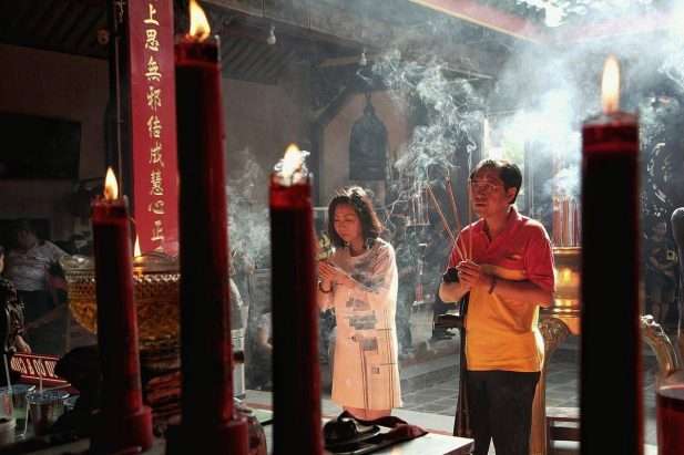 Cảnh quay cận cảnh tập trung vào một người phụ nữ và một người đàn ông đang chắp tay cầu nguyện trước một bàn thờ trong một ngôi chùa với khói hương bốc lên.